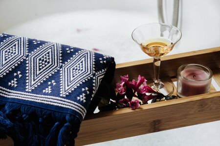 wooden shelf on the bathtub with glass of white wine, pink flowers and natural bath accessories