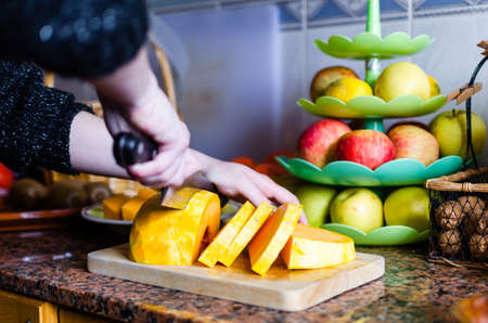 Beautiful caucasian girl with a large knife slicing a pumpkin on top of a wooden chopper / board on a brown marble countertop with black dots and a fruit bowl full of apples in the background with a blue and white tile wall.の写真素材