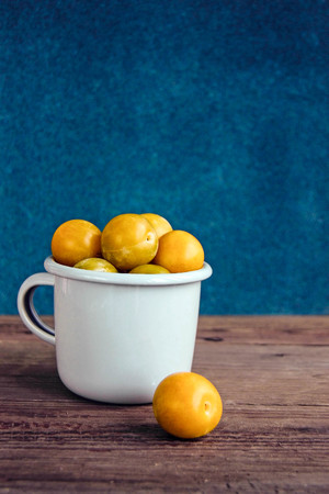 Fresh yellow plums in enameled cup on wooden rustic background and blue velvet wall. Russian homemade fruit. Front view. Copy spaceの写真素材