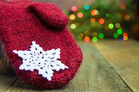 Red wool mitten with white cotton crocheted snowflake on the rustic wooden background. Red, blue, yellow and green spots of christmas lights. on fur tree. Card, postcard concept. Front view.Bokeh.の写真素材