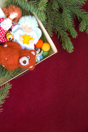 A wooden box with handmade Christmas toys: angel, bear, bee, doll near the branches of spruce on the burgundy velvet paper background. Red pear or jester red colors concept.の写真素材