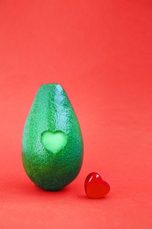 Fresh avocado with red glass heart on bright background. Minimal romantic love creative vegetarian concept for Valentine's day, vegan restaurant, wedding.の写真素材