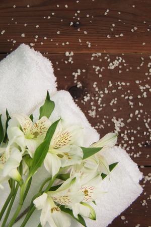 Beautiful alstroemeria flower and three rolls of clean white spa salon bath towels on brown wooden background. Top view flat lay copy space closeupの写真素材