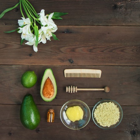Ingredients for vitamin avocado skin care facial mask green lime, bottle cosmetic oil,honey,dipper, oat-flakes, hair brush on brown wooden background. Alstroemeria flower. Top view close up copy spaceの写真素材