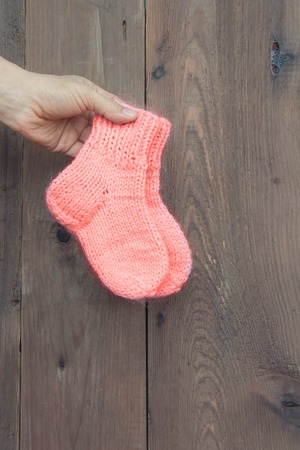 hands knitted baby socks color living coral in female hand on wooden wall background. Front...