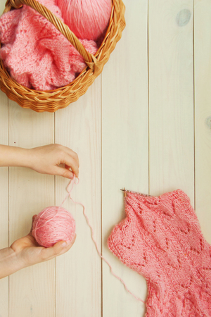 Hands with a coral wool thread on a ball in the process of knitting a karigan with an openwork pattern on wooden knitting needles and a basket on bright wooden rustic background. Flat lay top viewの写真素材