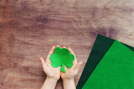 Child hands hold wool green clover, trefoil, shamrock for irish St. Patrick's day with sheets of felt on wooden background. Concept of DIY art project. Top view, copy spaceの写真素材