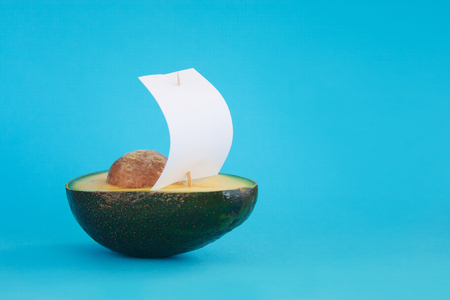 Beautiful creative decoration background idea with fresh green avocado yacht, sailboat with white sail on blue paper sea. Closeup. Front view. Copy space, vibrant food, freedom and adventure conceptの写真素材