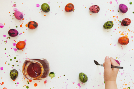 Hand child with a brush next to painted quail eggs with paints on the background of white paper with splashes and drops. Top view, flat lay. Copy space for text.の写真素材