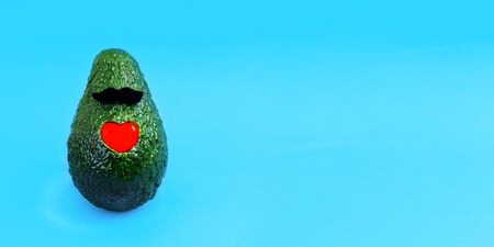 Fresh avocado with red glass heart and felt mustache on bright blue background. Minimal concept for holidays card, postcard. Web banner. Copy space for text. Front view.の写真素材