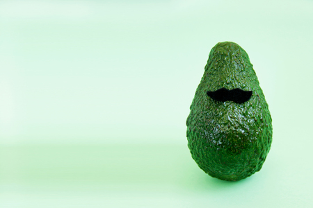 Fresh green avocado and black felt mustache on bright background. Minimal concept for holidays card, postcard. Web banner. Copy space for text. Front view.の写真素材