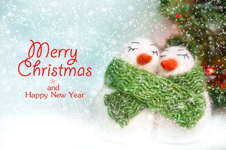 Family of white happy loving felted wool snowmen with green knitted scarf near Christmas tree with lights on bright blue background with snow. Front view. Christmas card concept with template.の写真素材