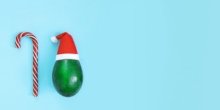 Fresh green avocado in a red hat next to a sweet candy cane on a bright blue background. Christmas concept, poster, tourist advertisements. Top view, web banner, copy spaceの写真素材