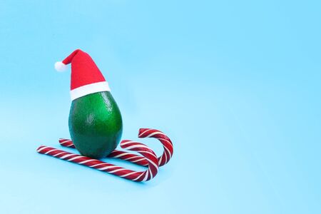 Christmas decoration idea with fresh green avocado skiing in santa hat on candy canes on bright blue background. Card concept. Closeup. Front view. Copy spaceの写真素材