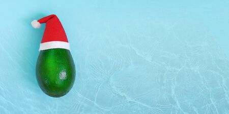 Fresh green avocado in a red and white Santa Claus hat. Christmas card concept. Water texture. Closeup. Top view. Copy spaceの写真素材