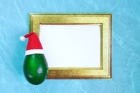 Fresh ripe green avocado in a red and white Santa Claus hat on a golden wooden frame with an empty place for the inscription. Water texture, tropical Christmas card concept. Flatlay, top view.の写真素材