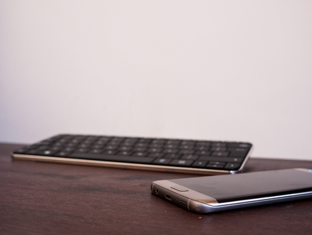 A smartphone lies on a wooden table in the foreground with a wireless keyboard in the background
