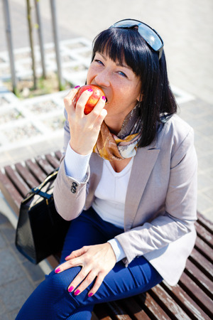 Stylish brunette woman looking at camera and eating an apple in the park, eating healthy and smilingの写真素材