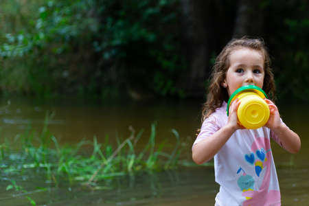 Little girl playing with some plastic buckets in the river. Girl with swimsuit and t-shirt. Children's photo in summer.の写真素材