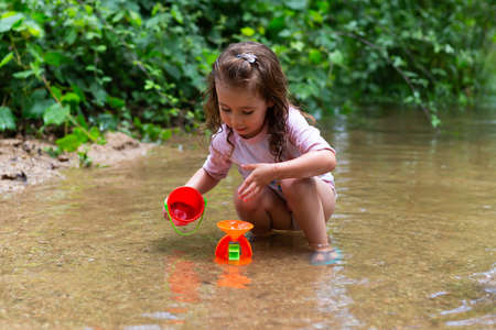 Little girl playing with some plastic buckets in the river. Girl with swimsuit and t-shirt. Children's photo in summer.の写真素材