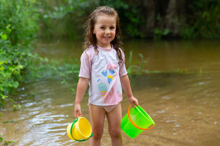 Little girl playing with some plastic buckets in the river. Girl with swimsuit and t-shirt. Children's photo in summer.の写真素材