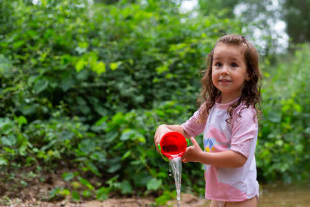 Little girl playing with some plastic buckets in the river. Girl with swimsuit and t-shirt. Children's photo in summer.の写真素材