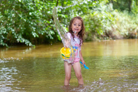 Little girl playing with some plastic buckets in the river. Girl with swimsuit and t-shirt. Children's photo in summer.の写真素材