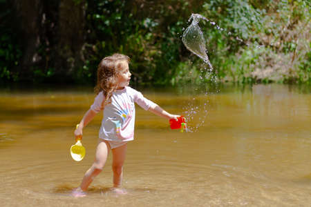Little girl playing with some plastic buckets in the river. Girl with swimsuit and t-shirt. Children's photo in summer.の写真素材