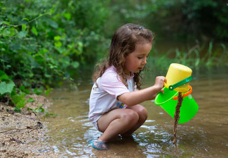 Little girl playing with some plastic buckets in the river. Girl with swimsuit and t-shirt. Children's photo in summer.の写真素材