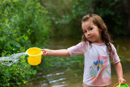 Little girl playing with some plastic buckets in the river. Girl with swimsuit and t-shirt. Children's photo in summer.の写真素材