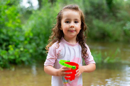 Little girl playing with some plastic buckets in the river. Girl with swimsuit and t-shirt. Children's photo in summer.の写真素材