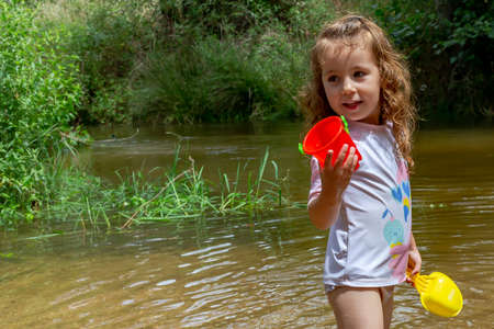 Little girl playing with some plastic buckets in the river. Girl with swimsuit and t-shirt. Children's photo in summer.の写真素材