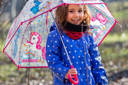 Caucasian blonde little girl in blue raincoat and unicorn umbrella looking at camera. Girl with umbrella outdoors. Girl enjoying a rainy day.の写真素材