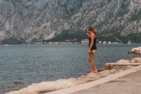 A young athletic woman stands on the shore and eats an apple in the summer .Kotor bay (Boka Kotorska), Montenegro, Europeの写真素材