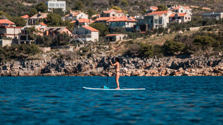 Young athletic woman paddling on a SUP stand up paddle board in Montenegro with a view of a beautiful village in the backgroundの写真素材