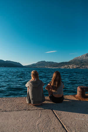 Back view: Two young women are sitting at the pier overlooking the sea in Montenegroの写真素材