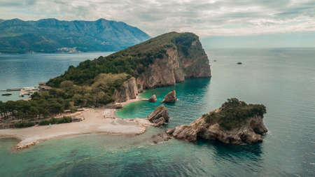 The beach and the cliff on the island of St. Nicholas in Budva with SUP stand up paddlers in water, Montenegro. Paradise beach on an island in the seaの写真素材