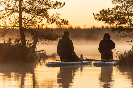 Young couple sitting on SUP stand up paddle board and drink tea  at sunrise in swamp. Early summer morning activityの写真素材