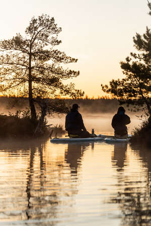 Young couple sitting on SUP stand up paddle board and drink tea  at sunrise in swamp. Early summer morning activityの写真素材