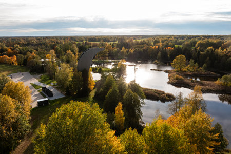 A curved observation tower rises above vibrant autumn trees, with a serene body of water reflecting the sky. A parking area is visible to the left.の写真素材