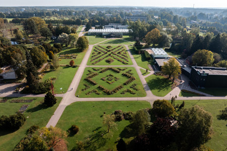 An aerial view of a botanical garden features geometric flower beds, pathways, and greenhouses, surrounded by lush greenery and urban buildings in the distance.の写真素材