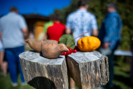 A wooden bird and colorful gourds sit on a rustic table. People gather in the background, with greenery and a clear blue sky suggesting a sunny day.の写真素材