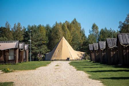 A large beige teepee tent stands at the end of a gravel path, surrounded by wooden cabins with...
