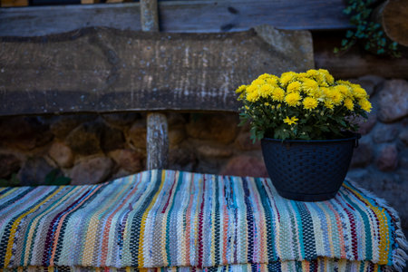 A rustic wooden bench is set against a stone wall, adorned with a striped woven blanket. A black pot with vibrant yellow flowers adds color to the scene.の写真素材