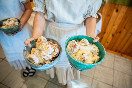 Two individuals in traditional clothing hold green ceramic bowls with desserts, possibly dumplings, in a rustic setting with wooden paneling and tiled flooring.の写真素材