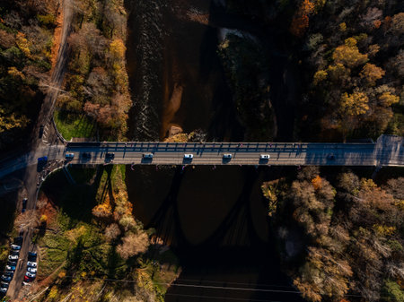 An aerial perspective of a bridge in Sigulda, Latvia, with vehicles crossing over a river. The scene is framed by a forest in vibrant autumn colors.の写真素材