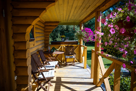A log cabin porch features rustic wooden chairs, a round table, and colorful hanging flower baskets, set against a lush green garden backdrop.の写真素材