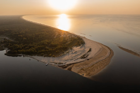 Aerial view of a narrow sandy peninsula extending into calm waters, surrounded by dense green forests, with the sun setting on the horizon.の写真素材