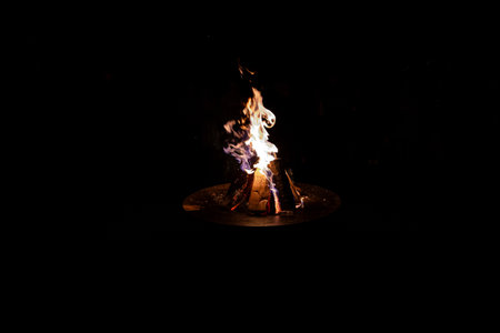 A campfire burns brightly in a circular metal fire pit, with logs in a teepee formation. The flames'...