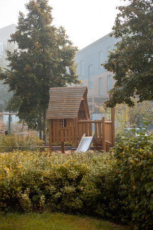 A wooden playhouse with a slide is surrounded by lush bushes and tall trees. A modern multi story building stands in the misty background, adding urban contrast.の写真素材